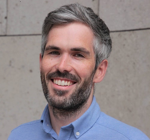 Coloured image of John Cooney looking at the camera smiling wearing a light blue collared shirt against an exterior walled background