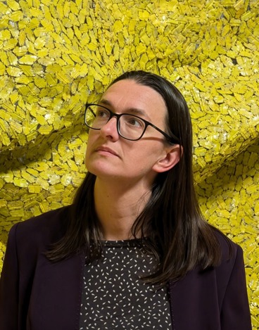 Image of Alex MacLaren looking away from the camera towards the top left of the image, wearing black framed glasses, a black top with white flecks, and a black jacket, standing against a yellow textured background