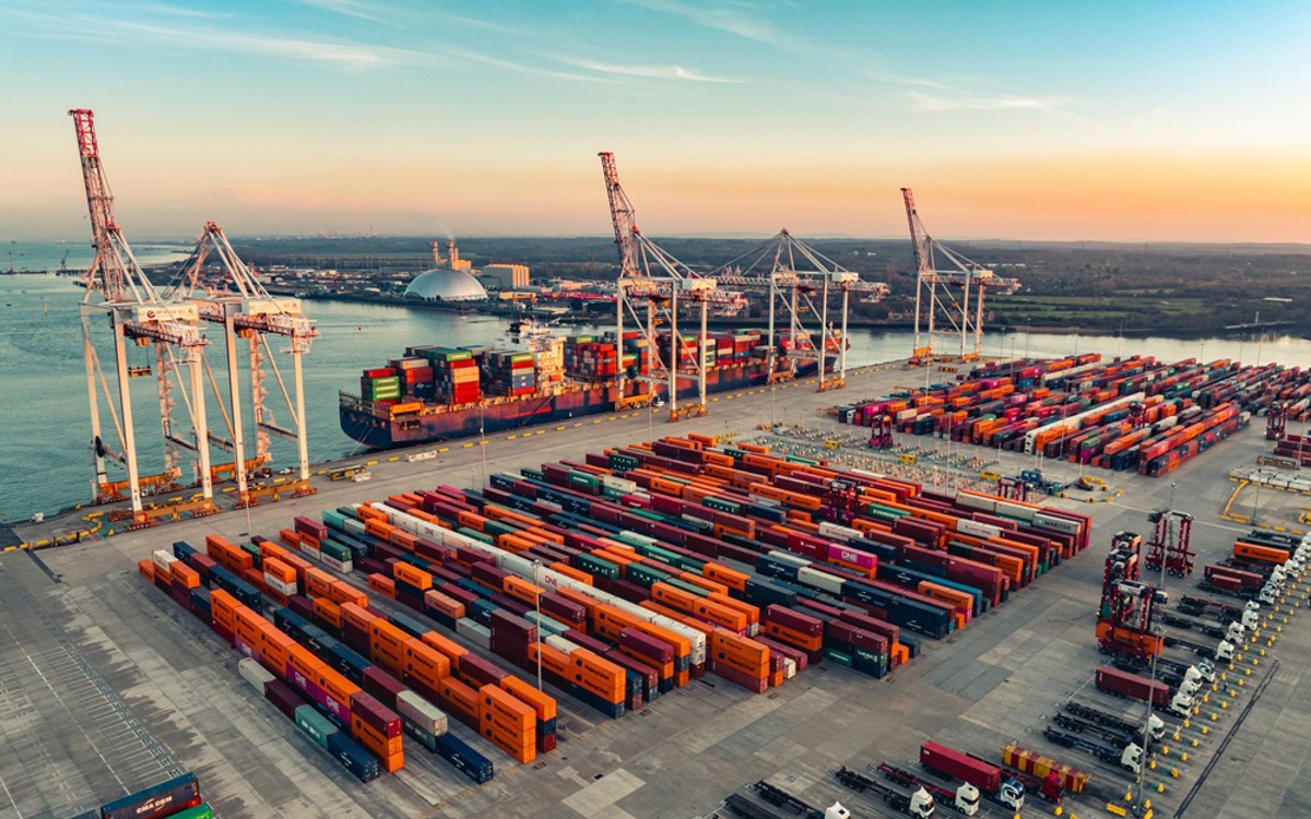 Shipping containers and cranes on a large harbour floor with dark grey sea and bright sunrise beyond.