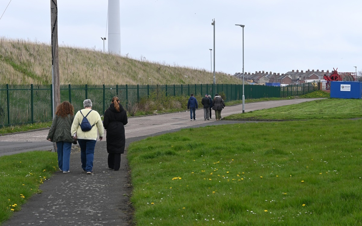 Small groups of people walking towards housing in the distance on a footpath with grass on the right and a fenced-off grassed area with wind turbines on the left.