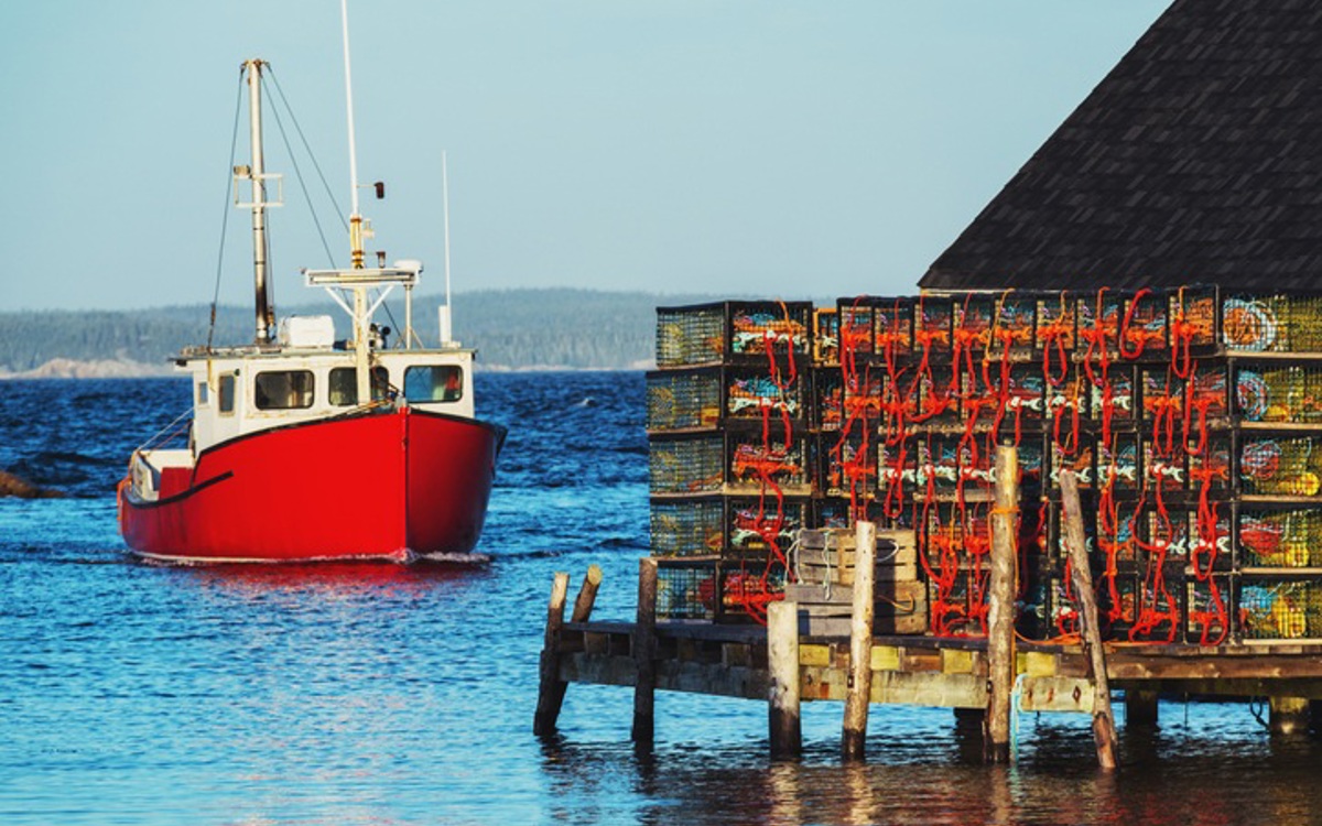 A red and white boat heading towards lobster pots stacked neatly on a wooden pier on a blue sea with grey-blue sky.