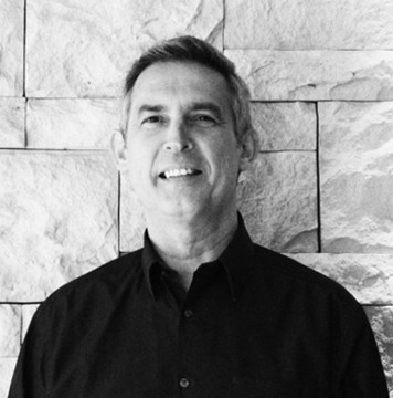 Black and white image of Kevin Murray looking into the camera smiling wearing a black collared shirt against a stone background