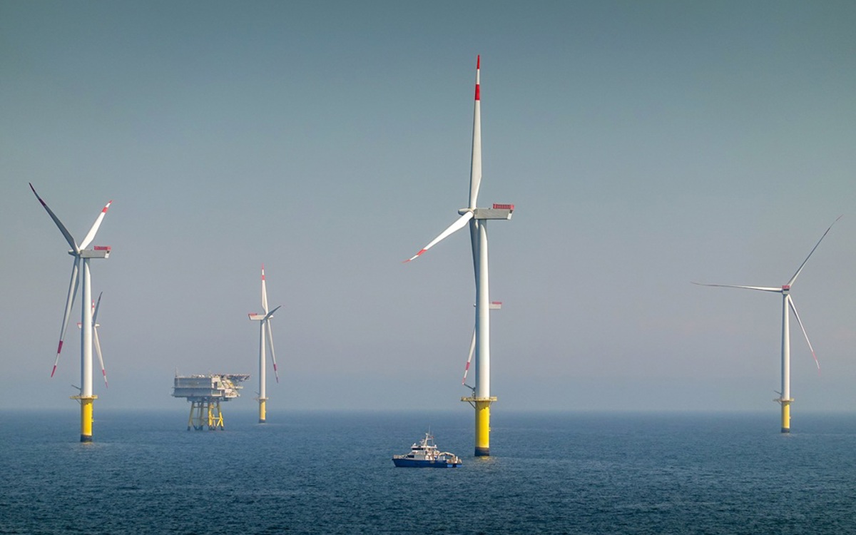 Four huge offshore wind turbines with white and red blades and white and yellow masts, as well as a substation, and a ship in the foreground that looks very small in comparison.