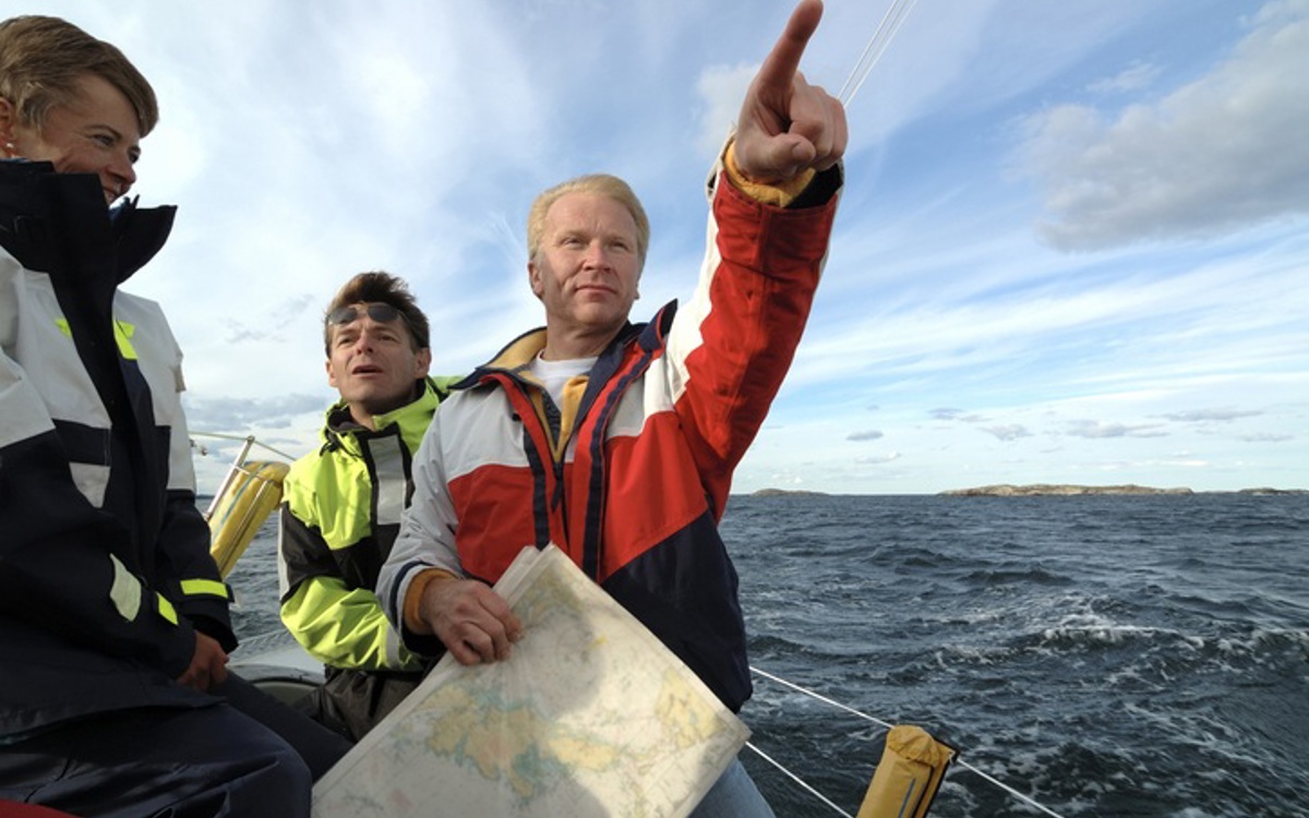 Three sailors standing on the edge of a boat on a dark grey sea with light blue and white partially cloudy sky, one sailor holds a chart and is pointing to the distance.