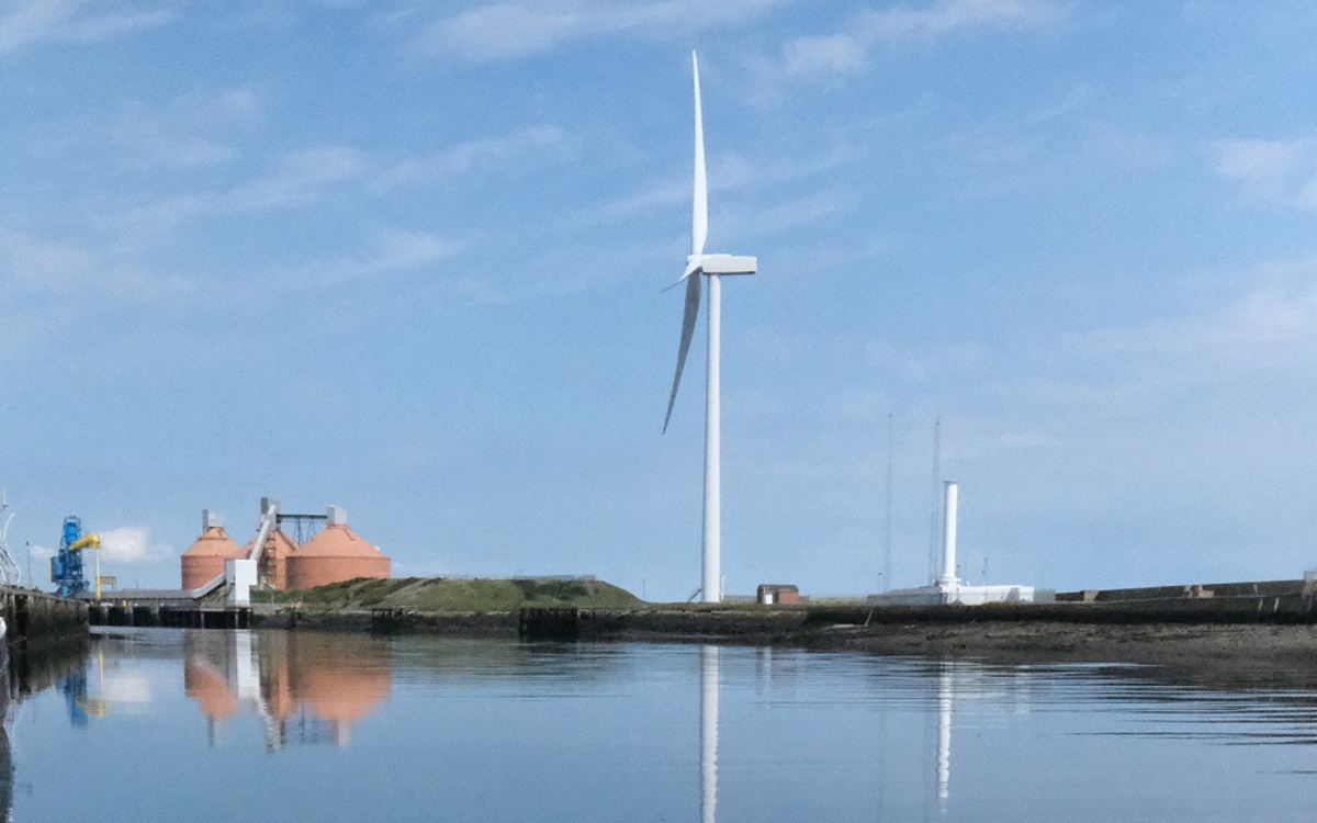 White wind turbine in the harbour beside the River Blyth, reflected in blue calm water with a blue and white partly cloudy sky.