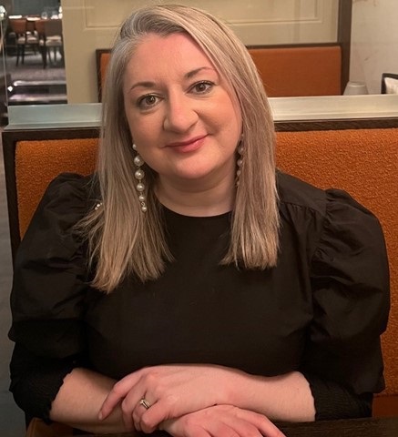 Coloured image of Maura McCormack sitting down at an orange coloured booth with a brown rim. Maura has her hands folded over her lap, is smiling at the camera, wearing a black long-sleeved top and dangly pearl shaped earings