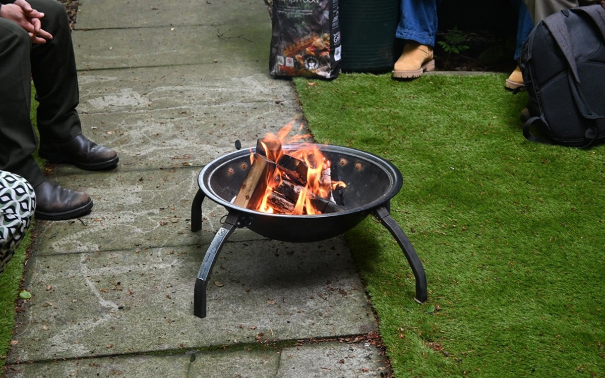 People seated around a wood fire burning in a round black raised firepit on a concrete paved footpath between artificial grass lawns